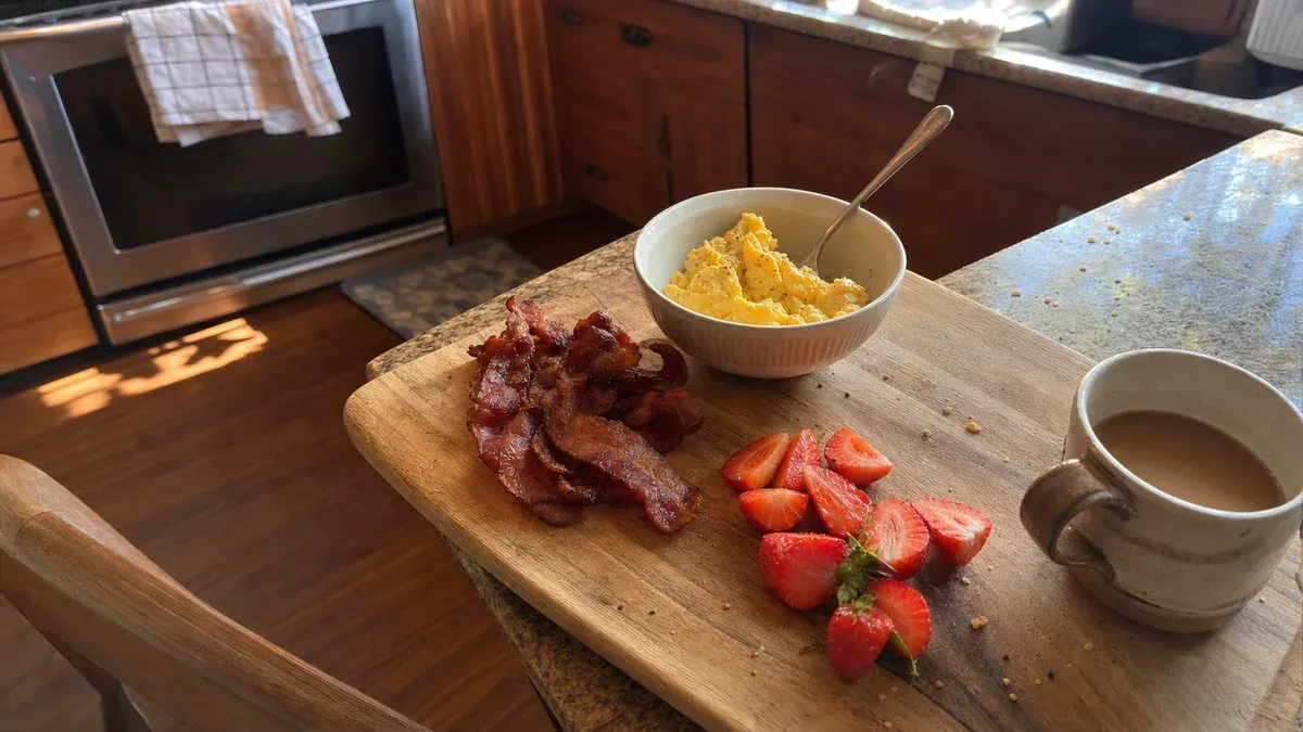 A casual breakfast board with crispy bacon, soft scrambled eggs, sliced strawberries, and a half-drunk cup of coffee on a wooden cutting board
