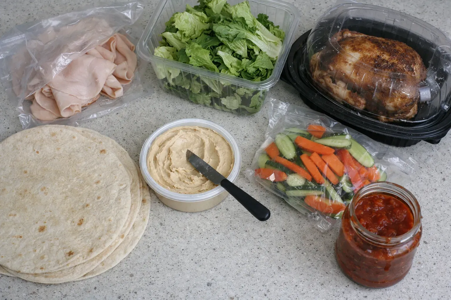 Simple no-cook ingredients including deli meat, tortillas, hummus, and rotisserie chicken on counter