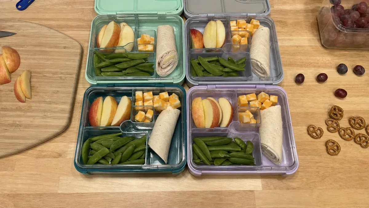 Overhead view of four open bento lunchboxes on a wooden counter being filled with cut fruit, cheese, wraps, and snap peas